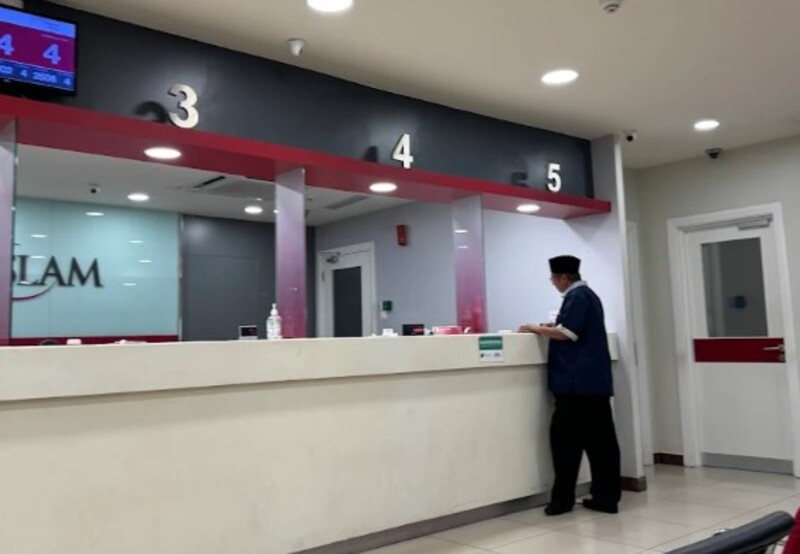 Pandangan dalaman cawangan Bank Islam yang menampilkan kaunter juruwang bernombor (3, 4, dan 5) dengan skema warna merah dan kelabu. Seorang lelaki berbaju biru dan bersongkok kelihatan di kaunter
