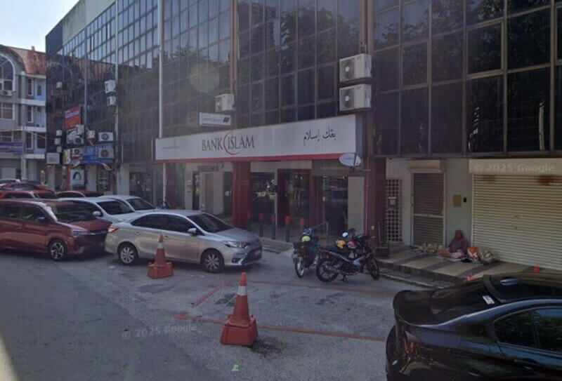 Pemandangan aras jalan cawangan Bank Islam Malaysia Berhad di Taman Melawati, memaparkan papan tanda kedai putih dengan aksen merah dan tulisan Jawi. Beberapa buah kereta dan motosikal diletakkan di hadapan bangunan berkaca itu